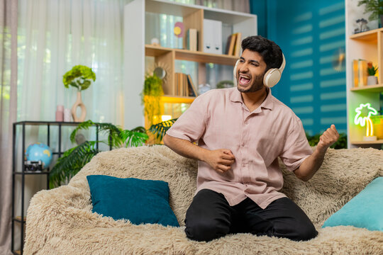Indian man on home sofa wears headphones while enjoying music with air guitar moves plus happy dance energy. Arabian guy on home sofa smiles during fun audio break with playful rhythm in living room.