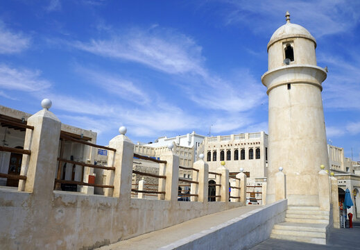 A cylindrical stone minaret with a domed cupola stands beside traditional vernacular buildings and a railed pedestrian ramp under a blue sky in Qatar.