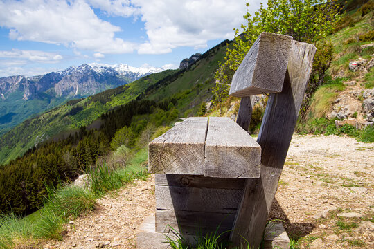 Side view of an empty weathered wooden bench on top of Monte Novegno mountain. Beautiful alpine summer landscape with green slopes and snow capped peaks under blue cloudy sky in Schio, Italy