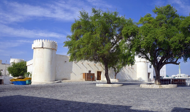 Lush green trees stand within a stone-paved courtyard before the whitewashed walls and crenellated round towers of Al Koot Fort beneath a blue sky in Doha, Qatar