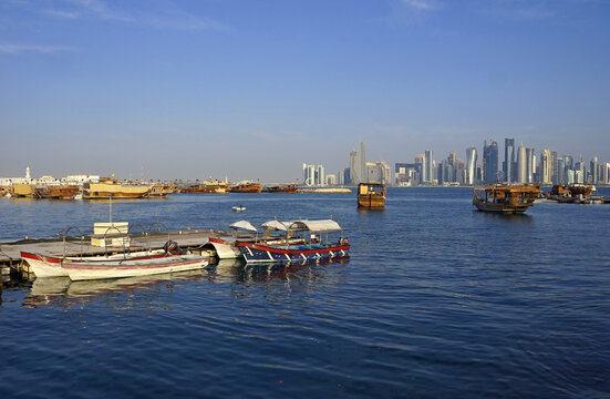 Small motorboats and traditional wooden dhows moor in Doha Bay with the modern West Bay skyline rising in the distance under a clear sky in Doha, Qatar