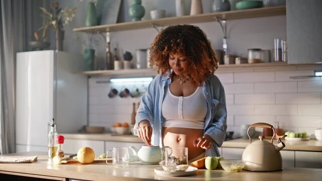 Pregnant african woman brewing herbal tea kitchen. Kettle pouring hot water.