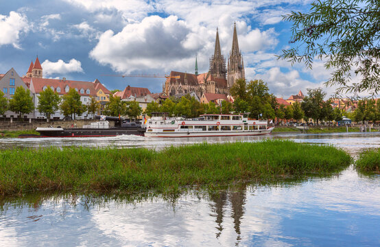 View across the Danube River toward Regensburg Cathedral, historic riverside buildings and passenger boats in Bavaria, Germany.