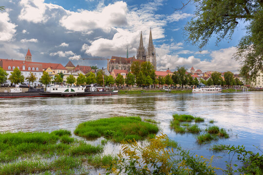 View across the Danube River toward Regensburg Cathedral, historic riverside buildings and passenger boats in Bavaria, Germany.