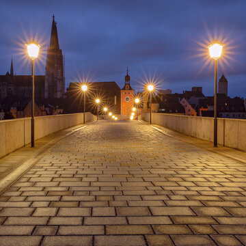 Illuminated Stone Bridge leading toward the historic center of Regensburg, Bavaria, Germany, during blue hour.