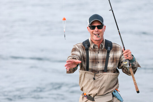 Man wearing plaid shirt and waders casts a fishing line into calm water, smiling under bright daylight while enjoying a cigar in Alaska&rsquo;s wilderness.