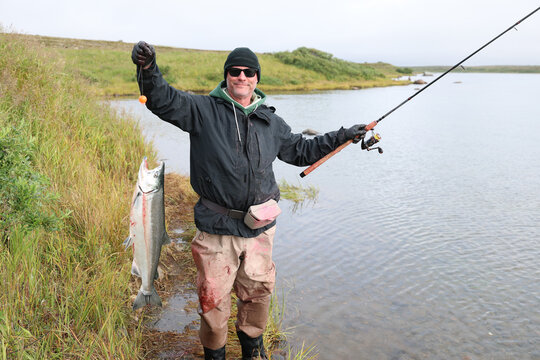 A proud man holds a large wild Coho salmon he caught on the Egegik River in Alaska, standing beside calm water in soft daylight that highlights the fish&rsquo;s silver body and the quiet wilderness around h