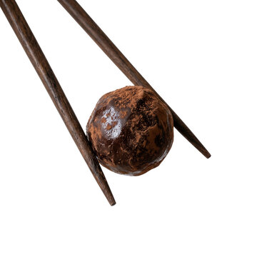 Una trufa de chocolate artesanal es sostenida delicadamente por un par de palillos de madera oscura sobre un fondo blanco puro, destacando su textura espolvoreada con cacao y su forma esf&eacute;rica