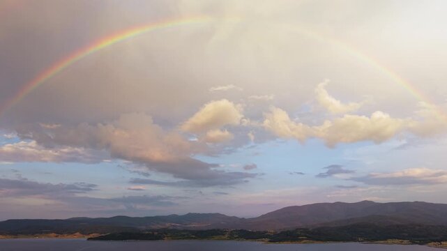 Drone view of Batak Reservoir in Bulgaria with rainbow arching over calm lake, rolling hills and distant mountains at golden hour