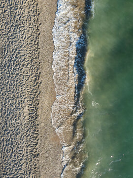 Vertical aerial drone shot directly above green ocean water washing onto a dark textured sand beach in straight line.