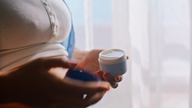 Cream jar pregnant belly closeup. Woman applying stretch mark moisturizer home.