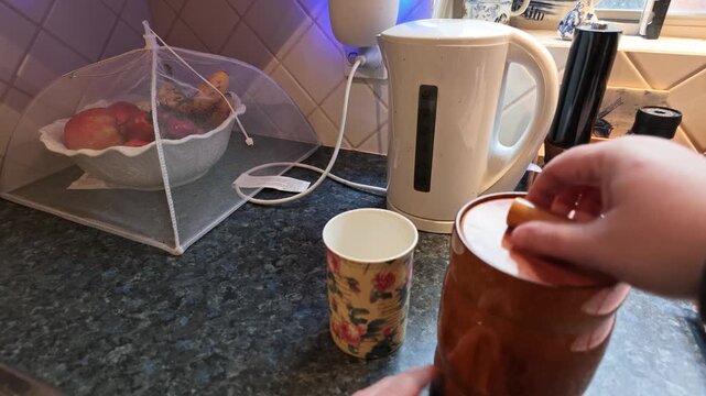 Human hand placing a floral mug on the kitchen counter, opening jar, taking a tea bag, and placing it into the cup