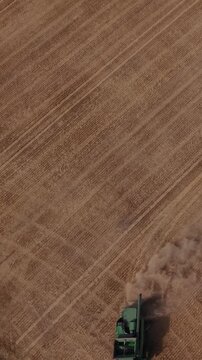 Vertical screen. Agricultural machinery operates in a golden field, harvesting crops while dust clouds rise, showcasing the expansive landscape under clear skies