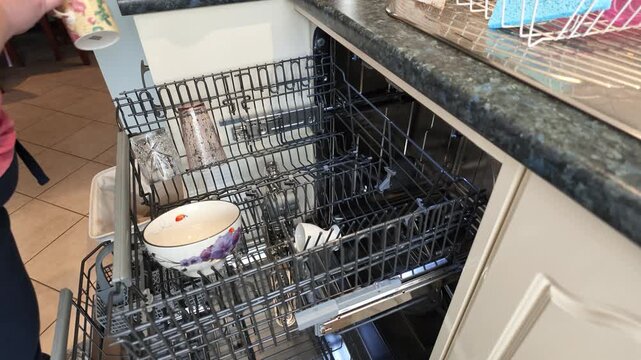 Person placing dirty bowls, glasses, and mugs onto the racks of a modern dishwasher before closing the door to begin the wash cycle