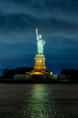 Statue of Liberty under storm clouds in New York at night. Centered vertical composition shows the copper figure, torch and pedestal above rippled water reflections. © white78