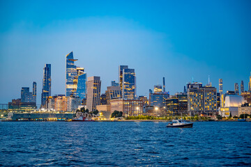 Hudson Yards skyline across the Hudson River at blue hour. Wide view shows illuminated towers, the Edge observation deck, waterfront buildings and a motorboat on rippled water. © white78