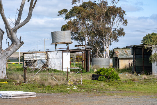 Dilapidated corrugated farm sheds, rusted wire fencing, and neglected elevated water tanks on a former farmyard or on an old rural homestead in regional Victoria, Australia.