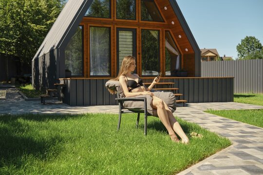 Young woman in bikini rests in a sun lounger in the backyard of a modern log house with smartphone