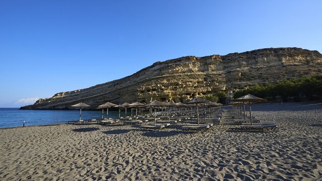Neolithic caves, former hippie paradise, empty beach with umbrellas and rocks under clear blue sky against calm sea, Matala, south coast, Crete, Greek Islands, Greece