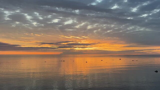 Wide panoramic sunset over the calm waters of the Baltic Sea with vibrant golden light breaking through storm clouds and birds flying over the coast in Gotland, Sweden.