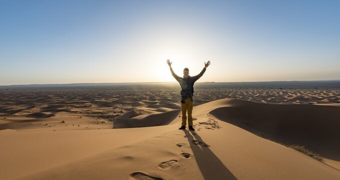 Young man stands on a sand dune and stretches his arms into the air, Erg Chebbi, Merzouga, Sahara, Morocco