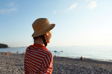 A young woman wearing a straw hat on a pebble beach looks at the sea during sunset. Her lifestyle reflects peace and tranquility in this summer vacation scenery by the ocean water coast.