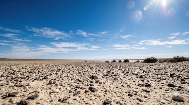 Low angle view of an arid desert landscape with a rocky gravel ground surface under a bright blue sky with scattered white clouds and a visible sun lens flare
