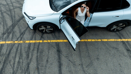 Woman stands near ev car. She poses by white ev car. Stylish ev car on road. © Nutchanart
