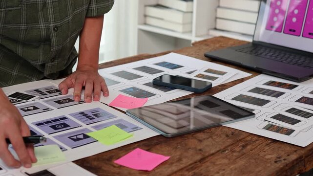 Creative web developer brainstorming and planning a new mobile application, working with ux wireframe and ui design templates on a wooden desk with a laptop and a tablet