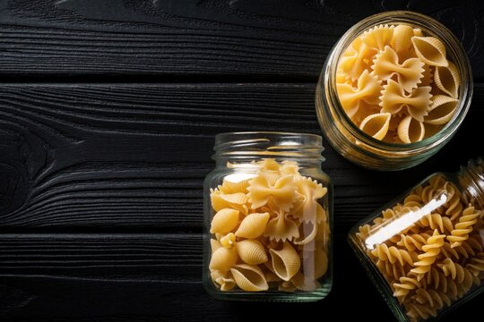 Glass jars filled with various dry pasta shapes like farfalle, conchiglie, and fusilli sit on a dark wooden table. Ingredients ready for cooking Italian meals.