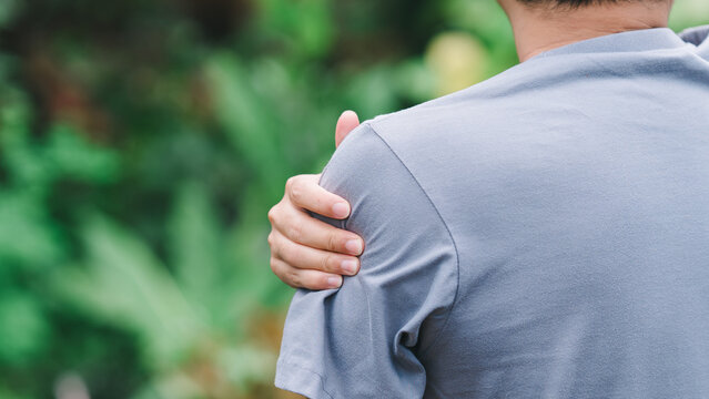 Close up rear view of man gripping his shoulder and deltoid muscle outdoors. Concept of sports injury, joint inflammation, physical therapy needs, and chronic arthritis body pain.