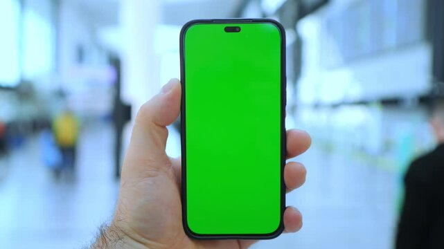 Hand holds phone with green screen. Background blurred public space. Three keyframes show same scene. Person stands in busy indoor area. Phone displays solid green color