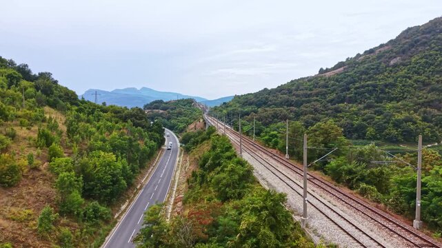 A view of railway tracks running parallel to a road in a rural area. Lush green hills are visible in the background under a cloudy sky. The scene shows the natural landscape in daylight.