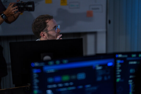A bespectacled male engineer working between bright monitoring screens scans the panel of charts while deciding what needs attention first inside a modern digital office
