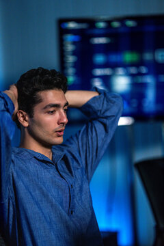A tired young male analyst rubbing his eyes in front of blue monitors rubs his eyes before returning to the incident queue in a night shift operations room in a modern technology company