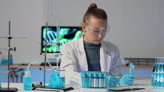 Young female scientist analyzing test tube samples and recording data in a modern genetics research laboratory with dna helix structure displayed on a computer screen