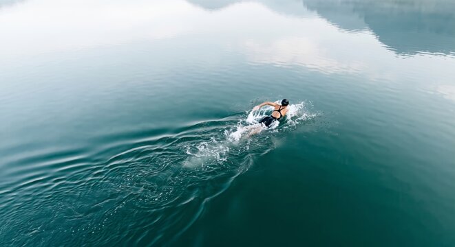 Female swimmer training in calm teal alpine lake water from high angle perspective creating white splashes and ripples while swimming freestyle in a vast scenic mountain landscape