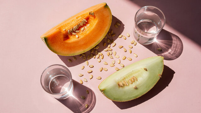 Fresh cantaloupe and honeydew melon slices with scattered seeds and glasses of water on a pink background, ideal for summer recipes, healthy diet promotion and beverage ads