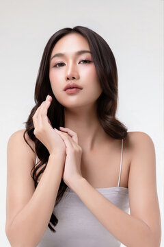 Beauty portrait of a young Asian woman with glowing, clear skin touching her face against a bright studio background, highlighting skincare and cosmetic beauty.