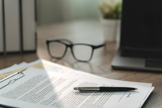 Pen resting across printed agreement documents on a clipboard beside eyeglasses and laptop, showing contract review, signature preparation and structured business documentation workflow.