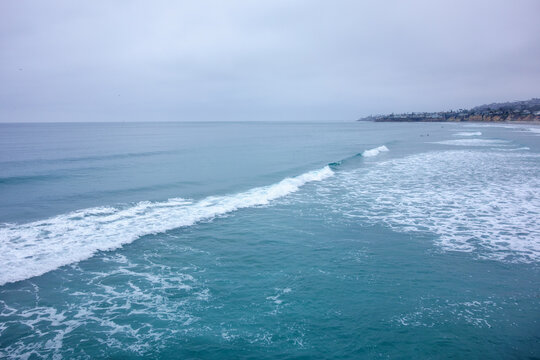A vast view of the turquoise blue Pacific ocean with white foaming waves rolling towards a distant cloudy coastline, San Diego, CA
