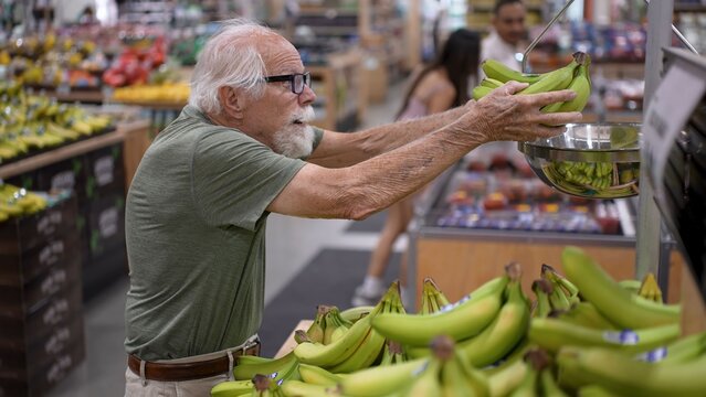 An elderly man is shopping in a grocery store, reaching for bananas in the fresh produce section. He is focused on his selection