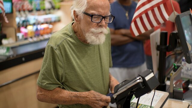An elderly man stands at the grocery store checkout counter and gets ready to pay for his items with a card while others wait
