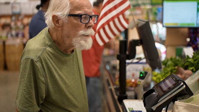 An elderly man stands at the checkout in a grocery store looking at the cashier while preparing to pay for his items