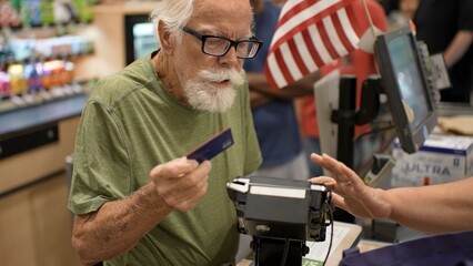 An elderly man stands at the grocery store checkout holding a card while preparing to pay for his items with staff assistance © Robert Peak