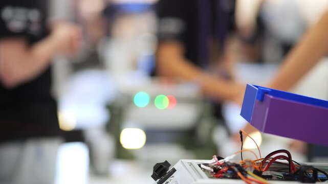 Close-up of robotic device with wires and glowing lights. Background shows blurred people at tech event. Focus on circuitry and electronics components