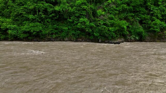 Mid level view across a wide muddy river rapidly flowing horizontally past a vibrant green forested bank in the Nepalese wilderness