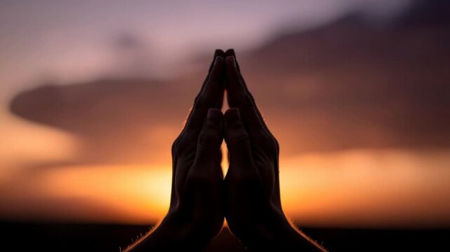Silhouette of hands pressed together in prayer with warm glowing sunset background