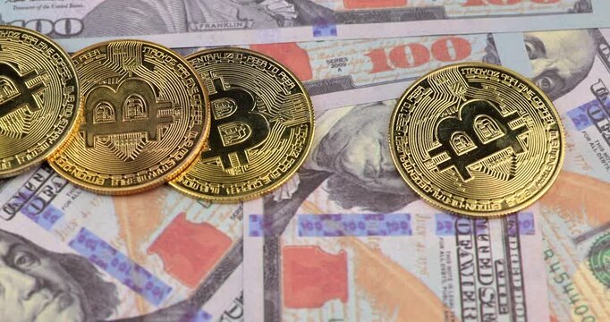 Horizontal close up of hand placing shiny physical golden bitcoin coins carefully on a spread stack of hundred US dollar banknotes in detailed perspective.