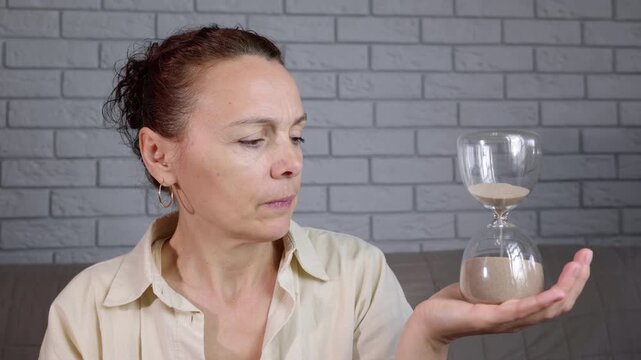 Pensive woman watching time pass in hourglass. Thoughtful middle-aged woman holding an hourglass and watching the sand run out, symbolizing the passage of time, aging, deadlines